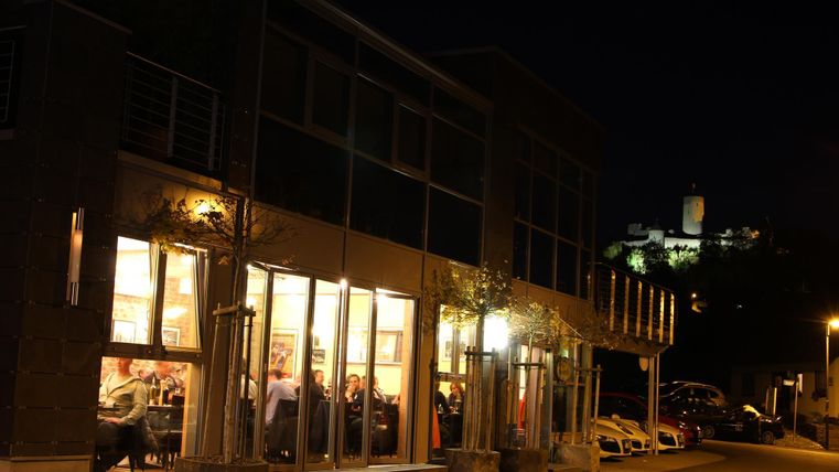 Ein modernes Restaurant mit beleuchteten Fenstern in der Nacht. Im Hintergrund ist eine erleuchtete Burg zu sehen.