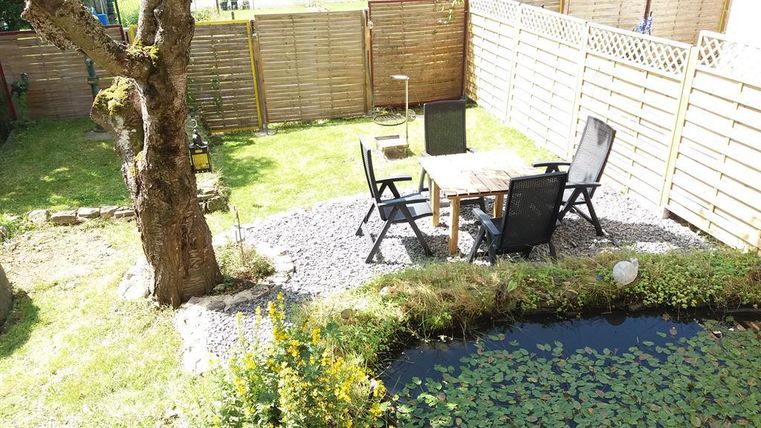 Ein schöner Garten mit einem Tisch und Stühlen auf Kies. Im Hintergrund befindet sich ein kleiner Teich mit Wasserpflanzen.