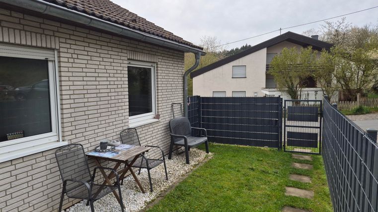 A cozy outdoor area with a table and chairs in front of a house. The green lawn and the fence create a peaceful atmosphere.