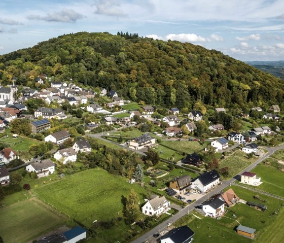 Luftbild von Aremberg: Ein Dorf mit weißen Häusern, grünen Wiesen und einem bewaldeten Hügel im Hintergrund unter blauem Himmel., © TI Hocheifel-Nürburgring©D.Ketz