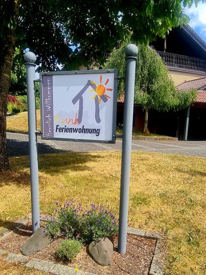 A sign for a holiday apartment stands in front of a building. The area is green and well-maintained, with some sunlight.