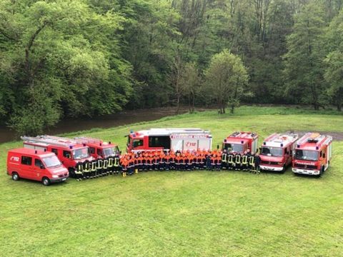 Eine Gruppe von Feuerwehrleuten steht in Uniform vor mehreren roten Feuerwehrfahrzeugen. Im Hintergrund sind grüne Bäume und ein ruhiger Fluss zu sehen.
