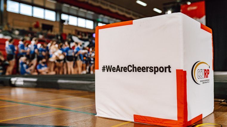 Een evenementenruimte met een opvallende witte kubus, die is gemarkeerd met de hashtag #WeAreCheersport. Op de achtergrond zijn veel cheerleading-atleten in blauwe uniformen te zien.
