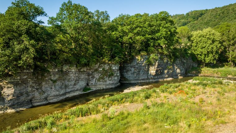 Ein ruhiger Fluss fließt zwischen bewaldeten Ufern und steilen Felsen. Die grüne Vegetation und der klare Himmel schaffen eine friedliche Landschaft.
