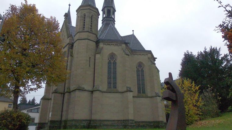 Eine historische Kirche mit spitzen Türmen und einer modernen Skulptur im Vordergrund. Der Himmel ist bewölkt und die Umgebung ist von herbstlichen Bäumen umgeben.