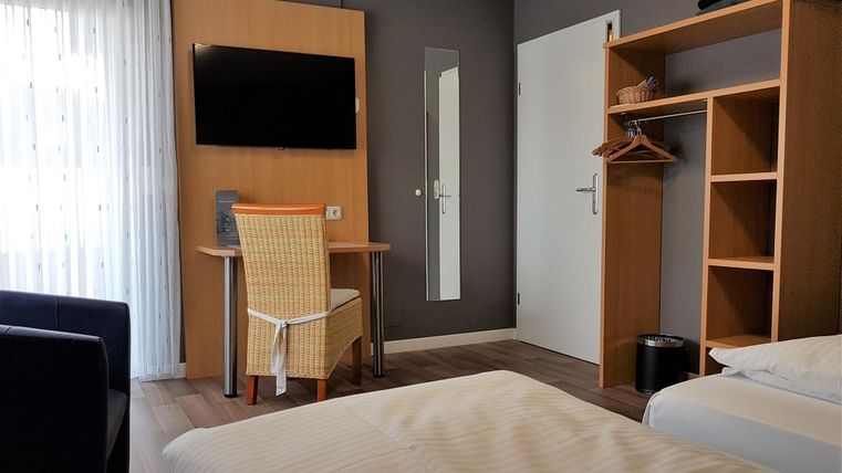 A modern hotel room with a double bed, a table, and a television. There is also a wardrobe and a wicker chair.