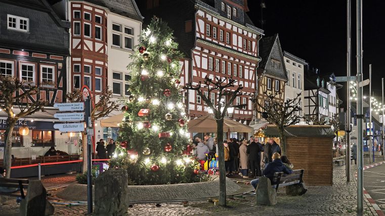 Eine festlich beleuchtete Straße mit einem großen Weihnachtsbaum. Menschen versammeln sich in der Abenddämmerung zwischen den hübsch dekorierten Gebäuden.