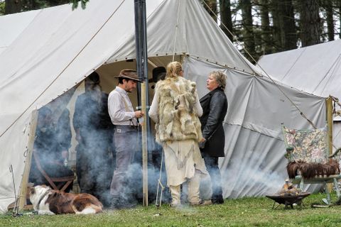 Ein Zeltlager in der Natur mit mehreren Personen, die sich unterhalten. Rauch zieht aus einem Lagertopf, während ein Hund im Gras entspannt.
