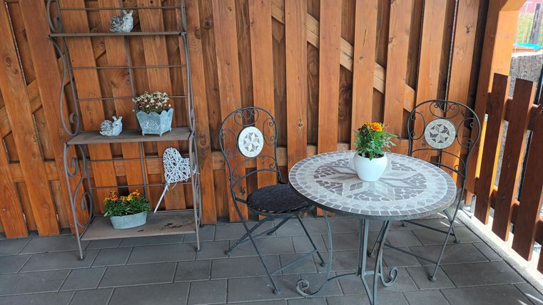 Een gezellige buitenruimte met een ronde tafel en twee stoelen. Op de achtergrond staat een rek met planten en decoratie.