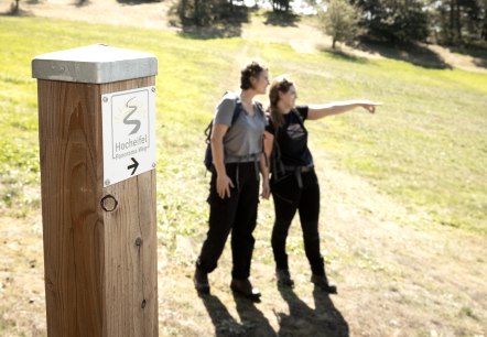 Zwei Wanderer stehen auf einem sonnigen Feldweg. Ein Holzpfosten zeigt den 'Hocheifel Panorama Weg' an. Eine Person zeigt in die Ferne., &copy; TI Hocheifel-N&uuml;rburgirng&copy;BaumannFotografie