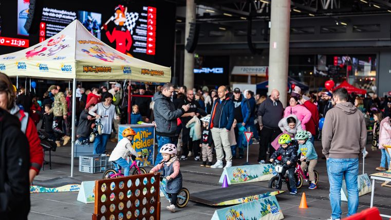 Eine lebhafte Veranstaltung mit vielen Menschen und Kindern, die auf kleinen Fahrrädern fahren. Im Hintergrund sind Zelte und eine Menge an Aktivitäten zu sehen.