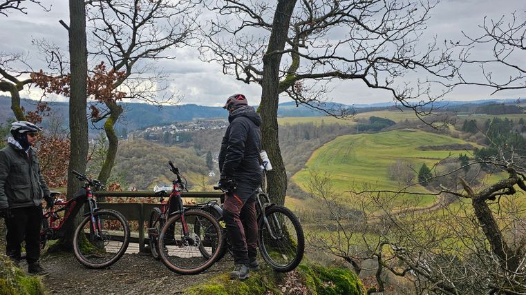 Zwei Mountainbiker stehen auf einem Aussichtspunkt in der Natur. Im Hintergrund ist eine grüne Landschaft mit Bäumen und Bergen zu sehen.