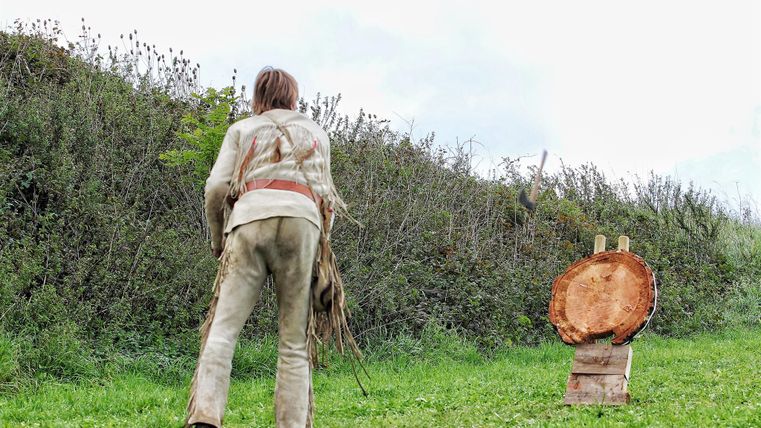 Eine Person steht im Freien und bereitet sich darauf vor, auf einen Holzscheit zu zielen. Im Hintergrund sind Sträucher und eine grüne Wiese sichtbar.