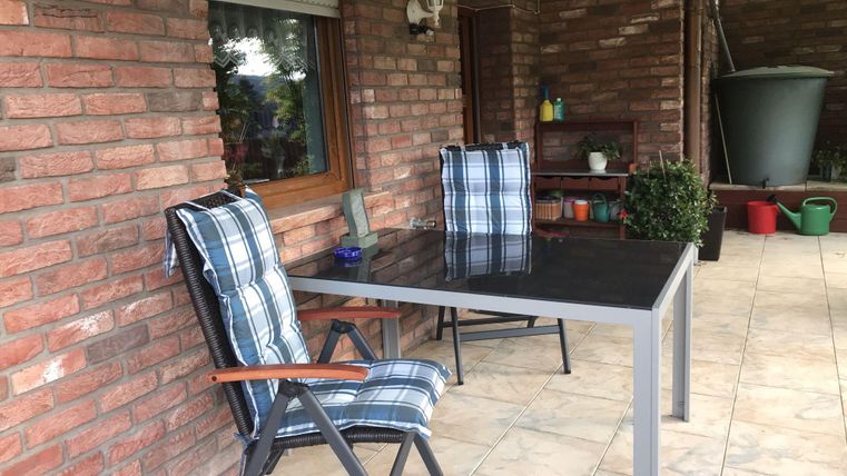 A cozy outdoor area with a table and two chairs. The walls are made of bricks, and there are plants in the background.