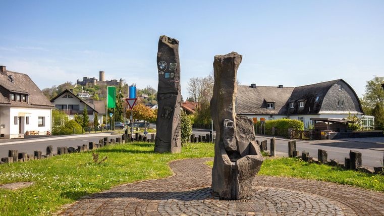 Een plaats met twee grote steinskulpturen en een groen grasveld. In de achtergrond zijn huizen en een heldere lucht te zien.