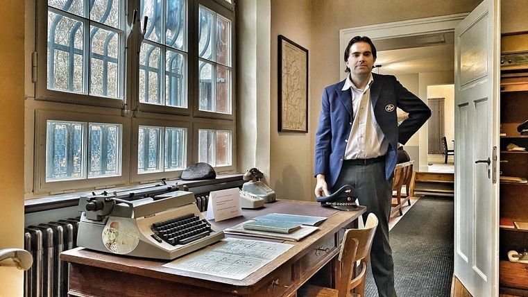 Een man in uniform staat in een werkkamer met bureau, typmachine en een groot raam. De kamer lijkt historisch ingericht.