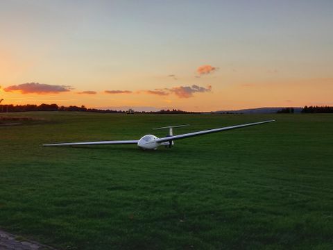 Ein Segelflugzeug steht auf einer grünen Wiese bei Sonnenuntergang. Der Himmel ist bunt und die Umgebung ist ruhig.