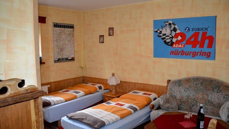 A simple room with two single beds and a coffee table. A poster with the Nürburgring motif hangs on the wall.