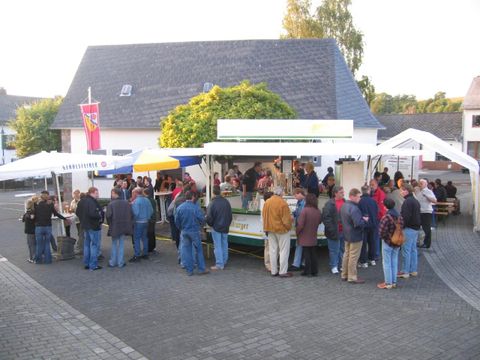 Ein belebter Marktstand mit vielen Menschen, die einkaufen. Im Hintergrund sind Zelte und eine entspannte Atmosphäre zu sehen.