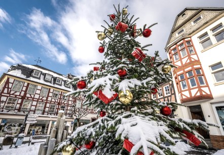 A festively decorated Christmas tree with red and gold baubles and presents, covered in snow, stands in front of half-timbered houses in Adenau., © VGVAdenau©Dominik_Ketz