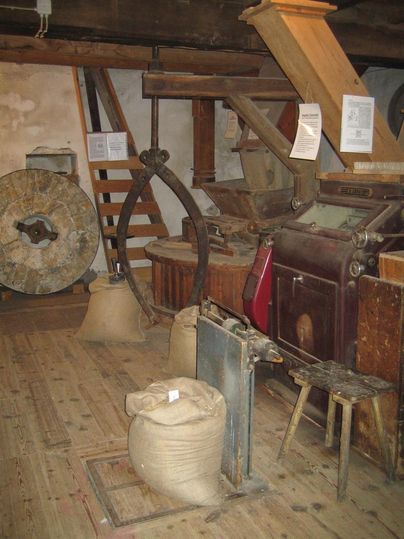 Ein historischer Mühlenraum mit einer Mühle, Säcken und verschiedenen Maschinen. Der Raum hat Holzböden und ist rustikal eingerichtet.
