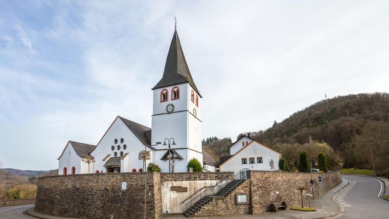 Eine Kirche mit spitzem Turm in einer kleinen Stadt. Im Hintergrund sind grüne Hügel und ein klarer blauer Himmel zu sehen.