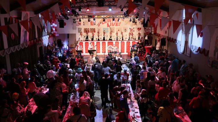 A lively event hall with many attendees dancing to music. Red and white decorations give the atmosphere a festive touch.