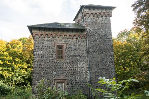 Ein alter Steinturm umgeben von Bäumen und Sträuchern. Die Fassade zeigt dekorative Elemente und Fenster.