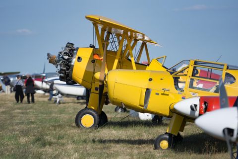 Ein gelbes Biplane steht auf einem Feld, umgeben von anderen Flugzeugen. Im Hintergrund sind Menschen zu sehen, die die Flugzeuge betrachten.