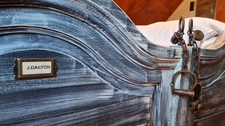 Een historisch bed met een kunstzinnig ontworpen hoofdeinde. De naam "J. Dalton" is aan de zijkant van het bed bevestigd.