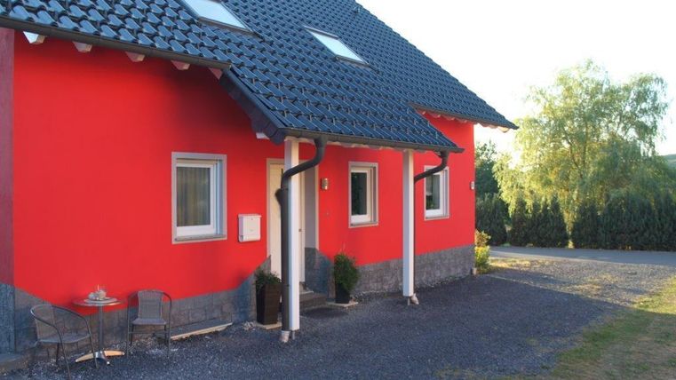 A red house with a gray roof and a small front porch. There are some plants and a cozy seating area next to it.
