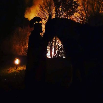 Eine Silhouette einer Person mit einem Pferd vor einem brennenden Hintergrund. Die Szene vermittelt eine mystische und dramatische Atmosphäre.