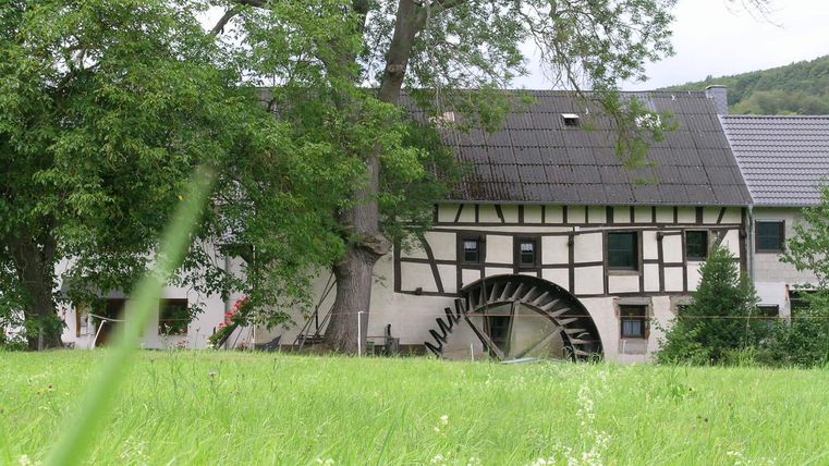 Fachwerkhaus mit Wasserrad und grüner Wiese im Vordergrund.