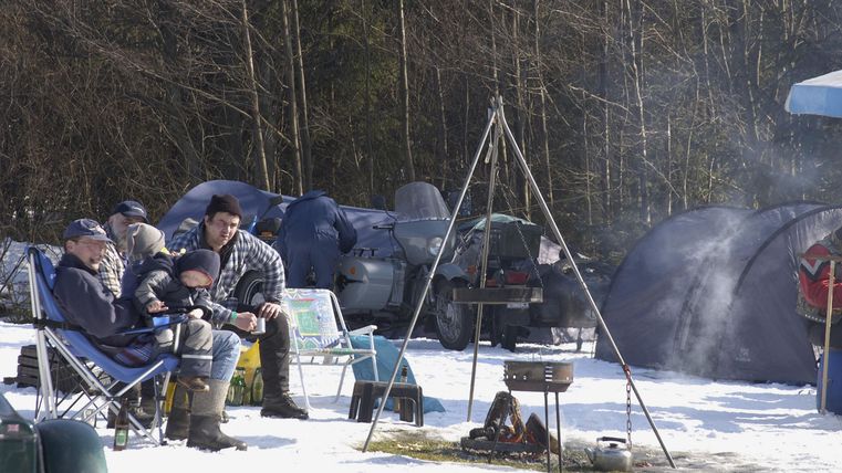 Family camping in the snow, surrounded by trees. A campfire burns in the foreground.