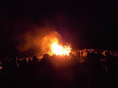 Ein großes Feuer brennt in der Dunkelheit, umgeben von einer Menschenmenge. Die Flammen leuchten hell und erzeugen eine festliche Atmosphäre.