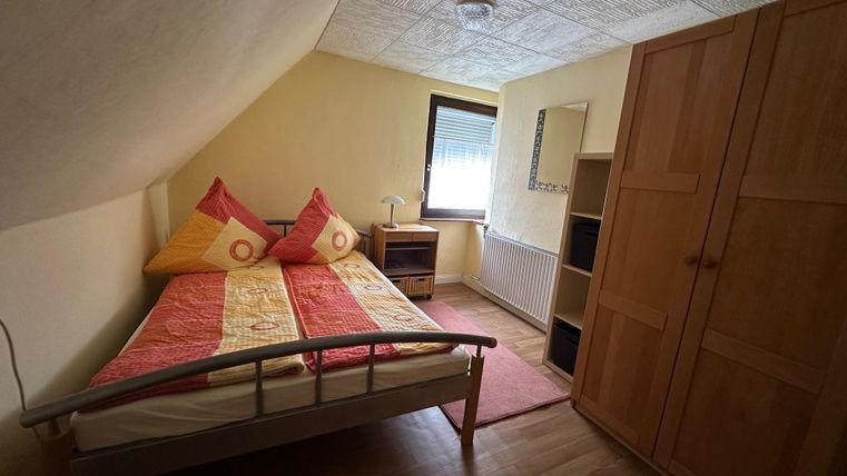 Een eenvoudige slaapkamer met twee eenpersoonsbedden, kussens in warme kleuren en een houten kast. De kamer heeft een lichte sfeer en een raam met uitzicht naar buiten.