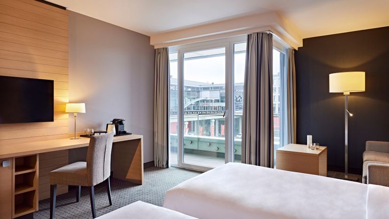 A modern hotel room with two beds, a desk, and a television. Large windows offer a view of the city.