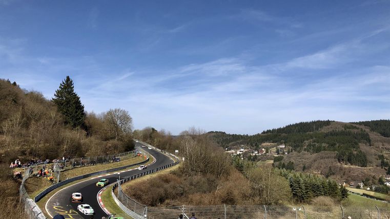 Une piste de course avec des sections sinueuses et plusieurs voitures, entourée de spectateurs. Le paysage est vallonné et boisé, sous un ciel bleu clair.