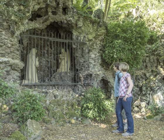 Zwei Personen stehen vor einer Felsengrotte mit Statuen, die durch ein Gitter geschützt sind, umgeben von grüner Vegetation., © VG Adenau©Kappest