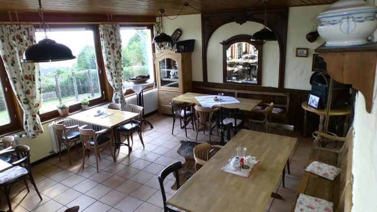 A cozy room with several wooden tables and chairs. Large windows provide a view of the outdoors and the walls are decorated with ornaments.