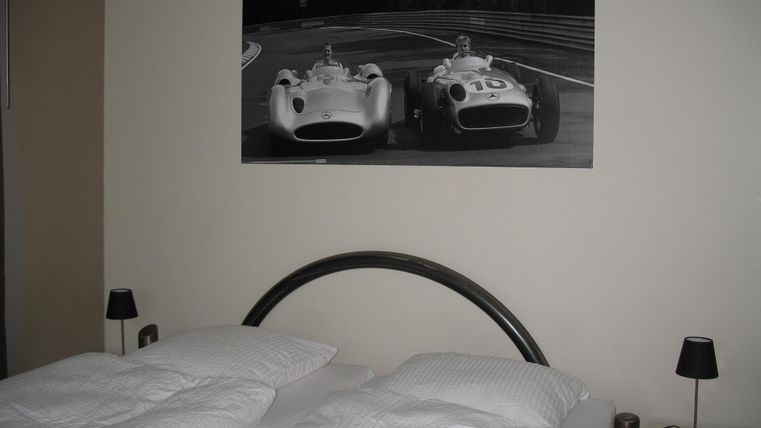 Ein modernes Schlafzimmer mit einem großen Bett und zwei Nachttischen. An der Wand hängt ein Schwarz-Weiß-Bild von klassischen Rennwagen.
