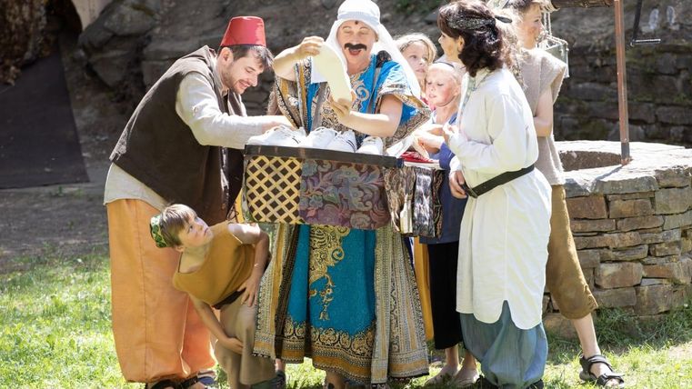 A group of children and adults in historical costumes gathers around a table. They appear to be having fun and experiencing something together.