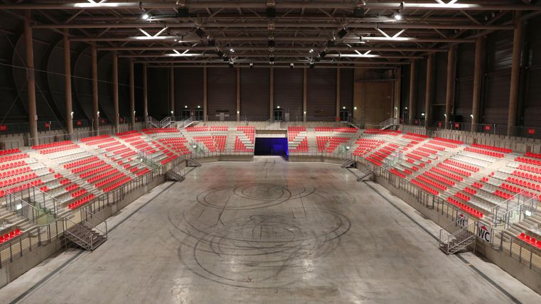 Een grote, lege hal met rode zitplaatsen en een gladde betonnen vloer. De muren zijn hoog en de verlichting is op de achtergrond zichtbaar.