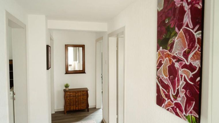 Ein heller Flur mit weißen Wänden und einem großen Spiegel. An der Wand hängt ein farbenfrohes Gemälde und es gibt eine kleine Holzkommode.
