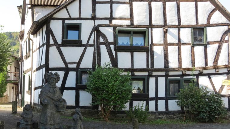 A traditional half-timbered house with white walls and visible wooden beams. In front of the house stands a statue with several figures.