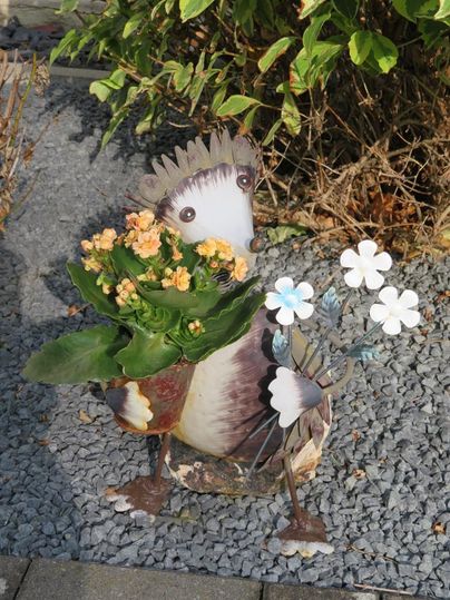 Une figurine décorative d'un animal tenant des fleurs et des plantes dans les mains. Elle se tient dans un espace jardin avec des cailloux et des plantes vertes en arrière-plan.