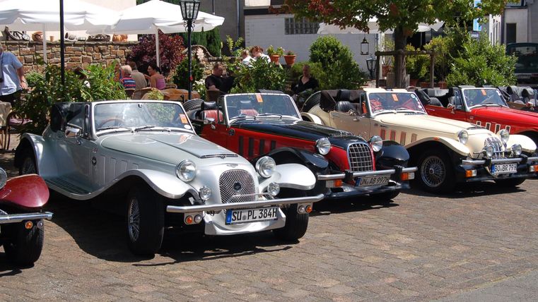Een reeks klassieke auto's staat geparkeerd op een stenen pad. Op de achtergrond zijn cafétafels en groene bomen te zien.