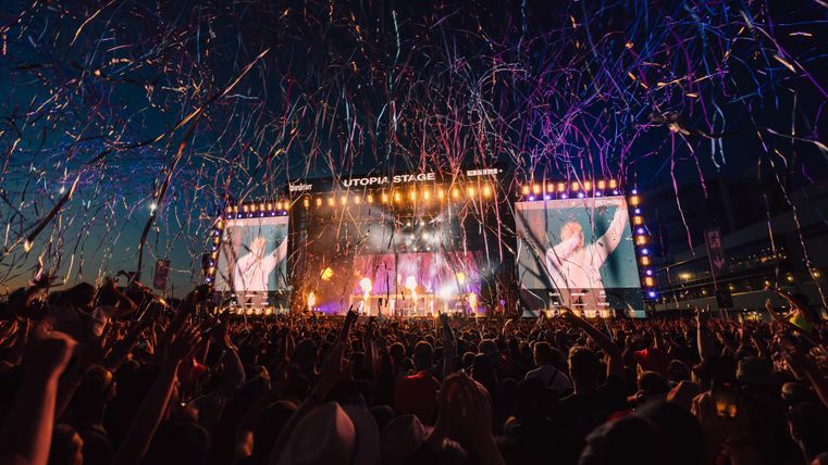 Ein lebhaftes Musikfestival mit einer großen Menschenmenge vor einer Bühne. Bunte Konfetti fliegen durch die Luft in einer festlichen Atmosphäre.