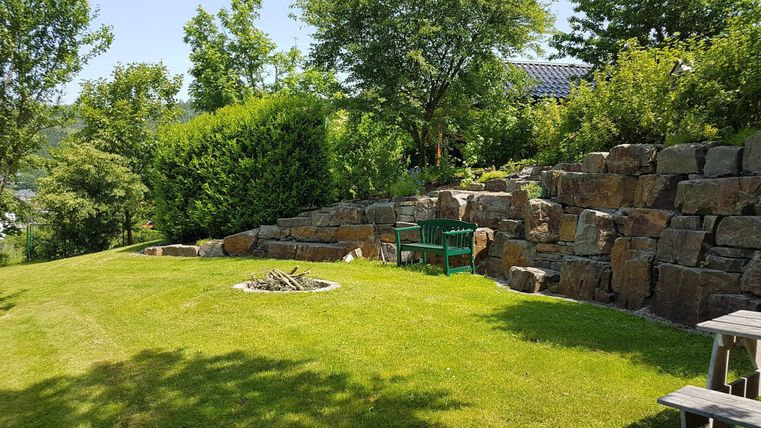 Eine gepflegte Gartenlandschaft mit Rasen und Felsen. Im Hintergrund steht eine grüne Bank und ein Feuerplatz.