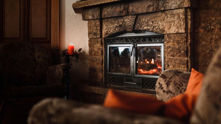 Een gezellige open haard met een stenen omhulsel en knapperend vuur. Op de voorgrond zijn een comfortabele bank en een kussen te zien.